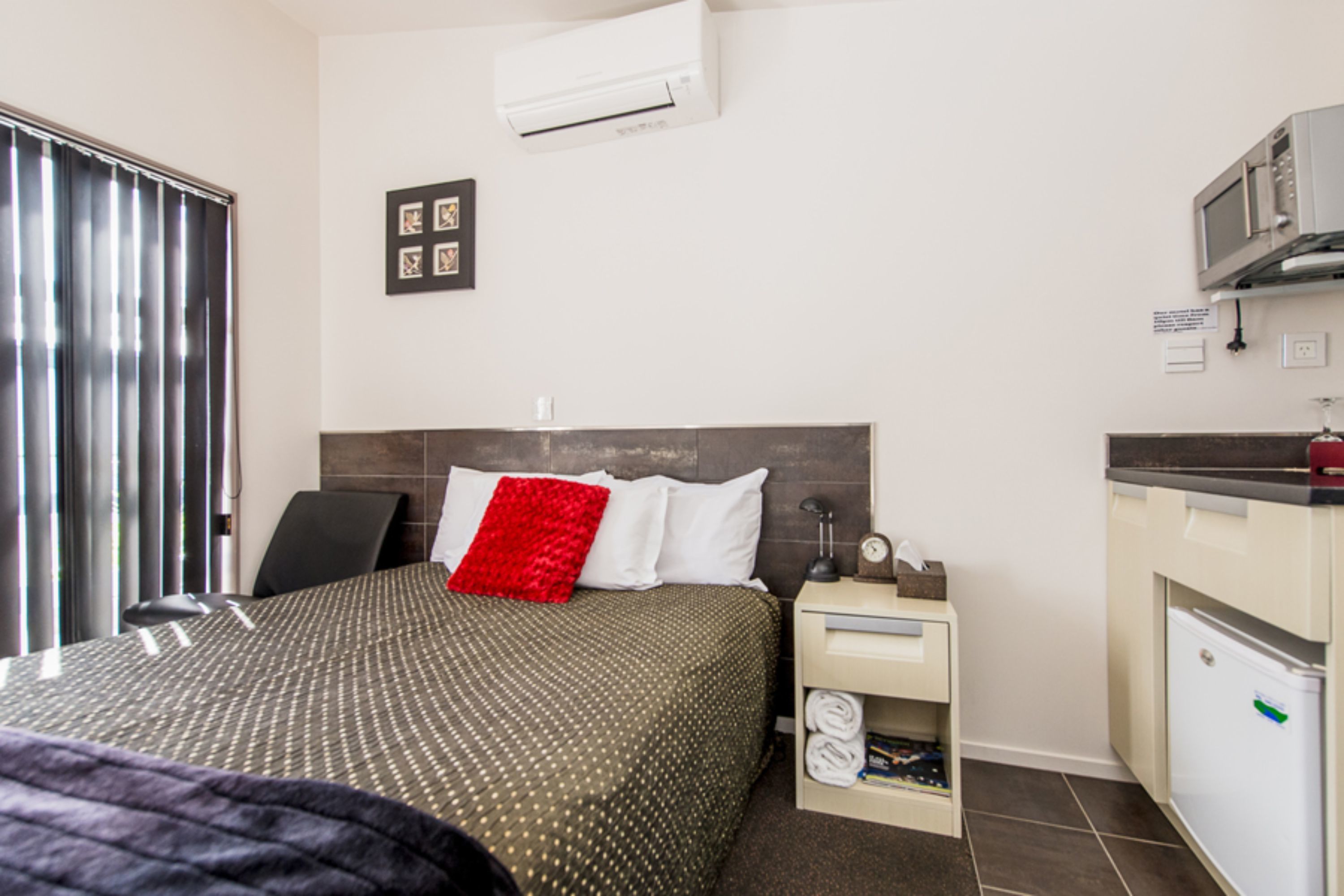 A hotel room with a bed, a desk, a chair, a small kitchenette, and a window with blinds.