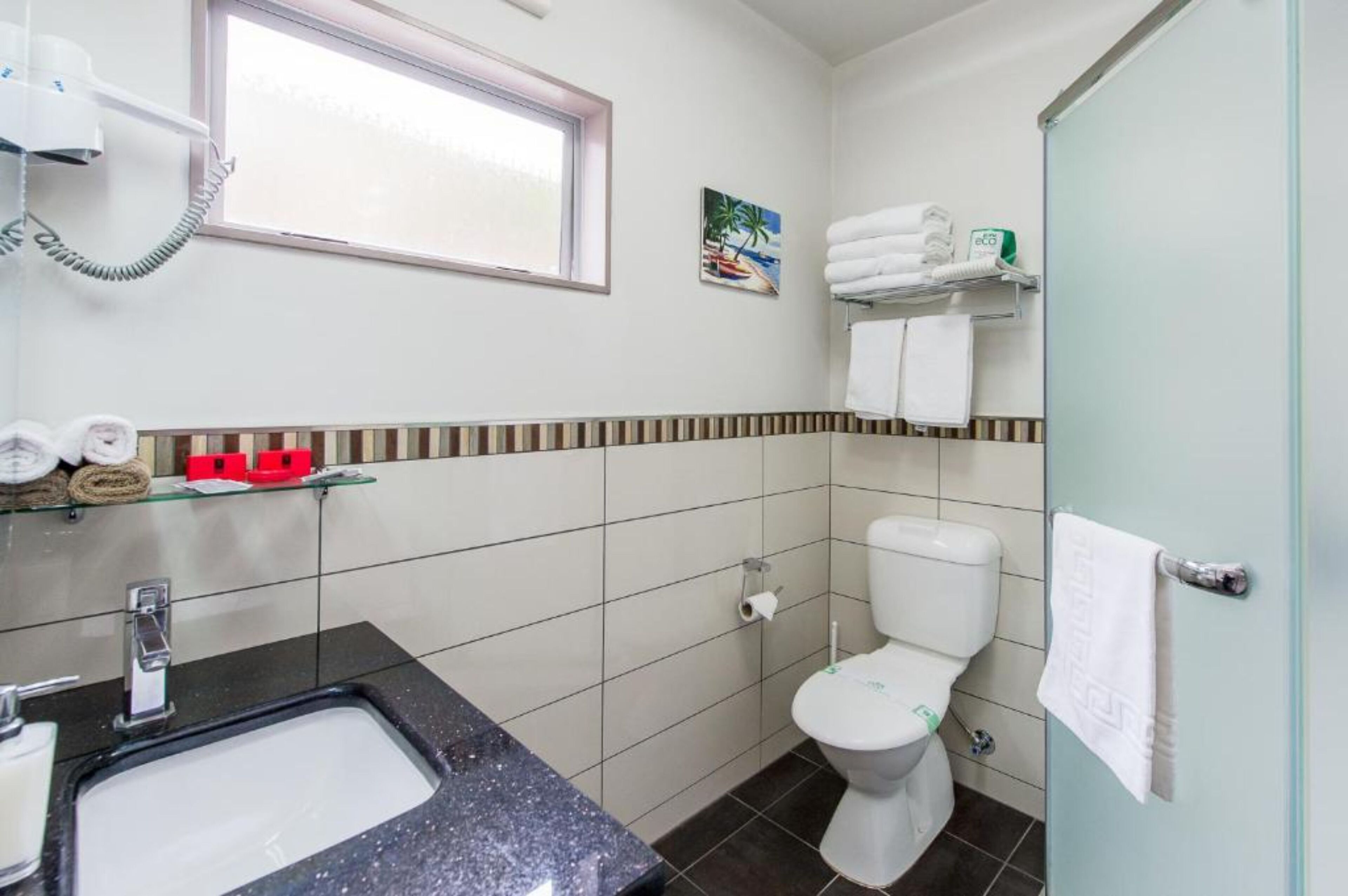 Casa de banho com lavatório, sanita e janela. Toalhas e produtos de higiene estão dispostos em prateleiras.