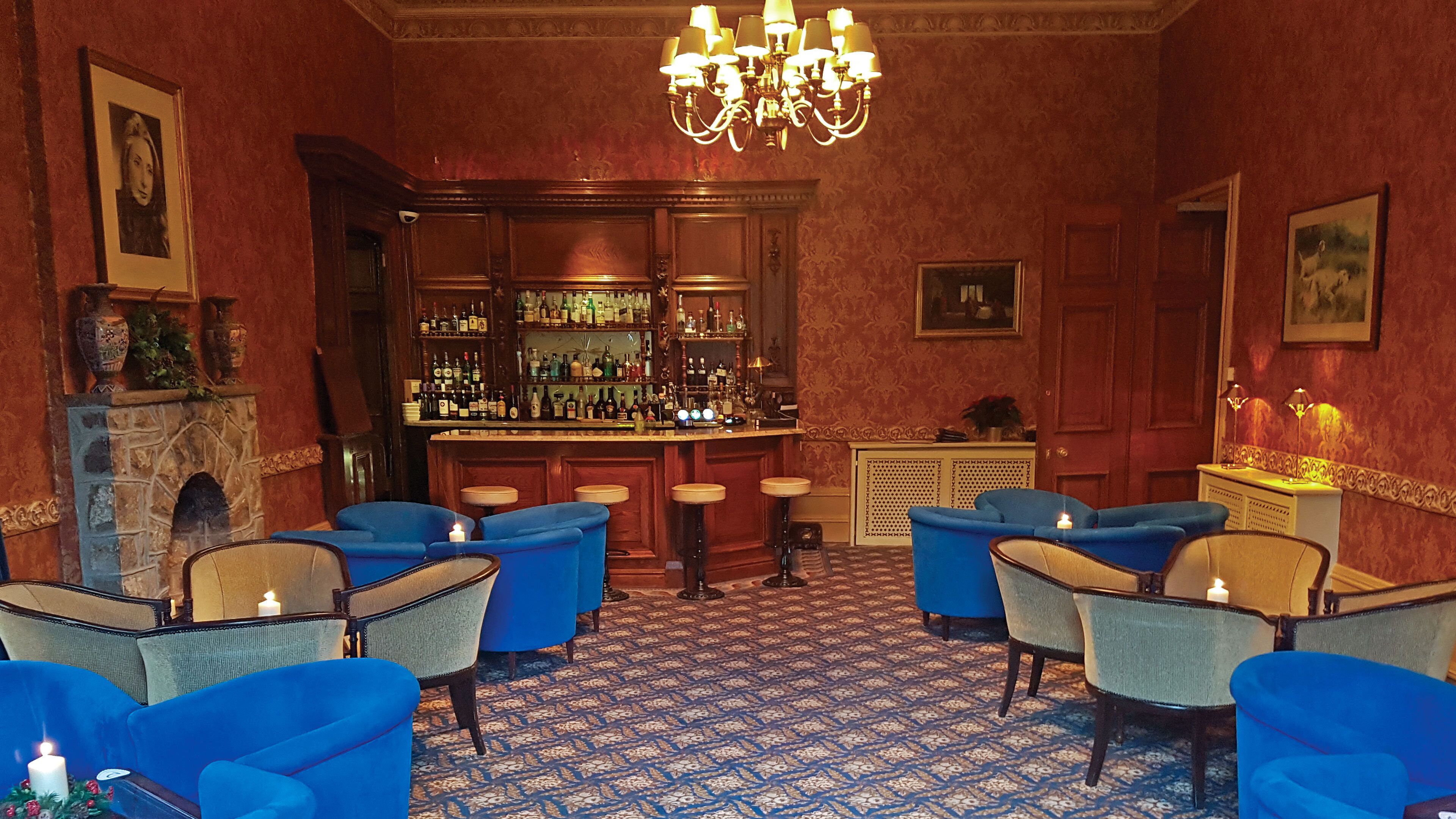 A bar area with a patterned carpet, blue upholstered chairs, a wooden bar counter with bottles, and a chandelier.