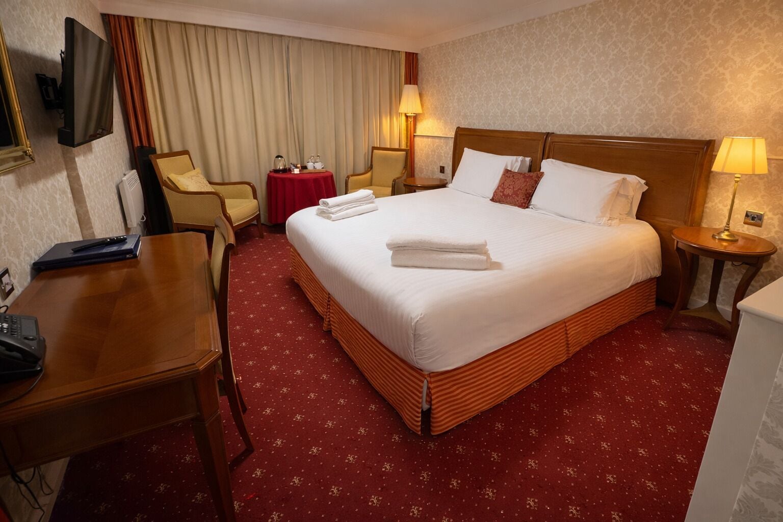 A hotel room with a large bed, a desk, a chair, a TV, and a table with a red tablecloth.