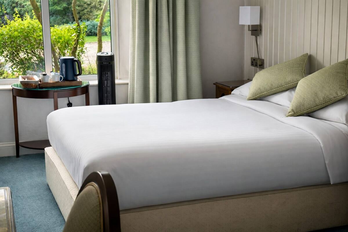 Een net opgemaakt bed met witte lakens, een nachtkastje met een lamp, en uitzicht op groen.
