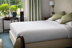 Una cama bien hecha con sábanas blancas, una mesita de noche con lámpara y vistas al exterior con vegetación.