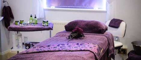 Un cuarto de spa con una camilla de masaje, una ventana con cortinas moradas y un estante con artículos de aseo.