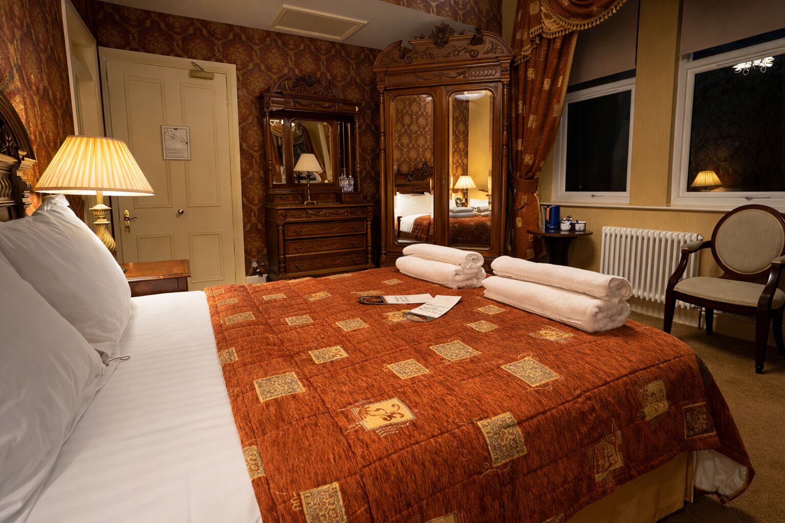 Una habitación de hotel con una cama grande, un armario de madera, un espejo y una ventana con cortinas.