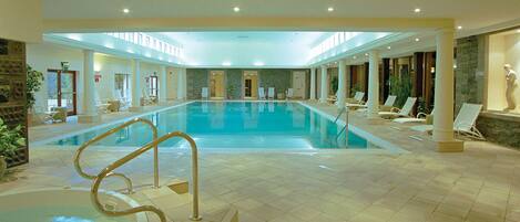 A large indoor swimming pool with a tiled floor, surrounded by lounge chairs and a stone wall.