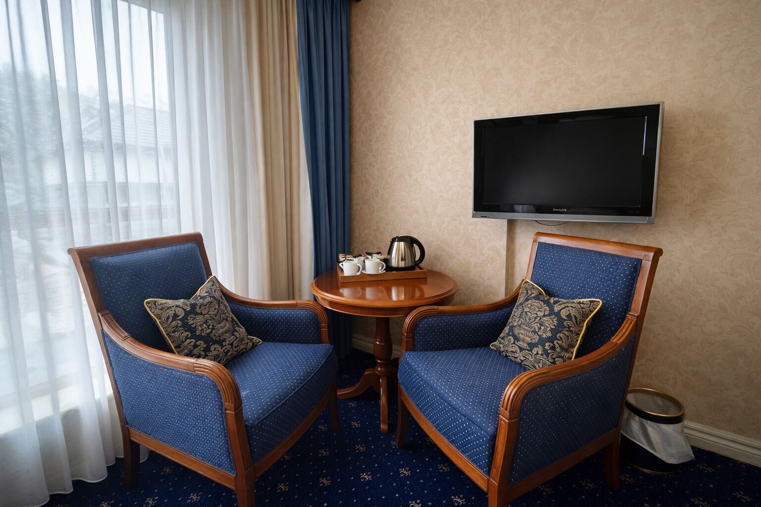 A hotel room with two blue upholstered chairs, a small round table with a coffee pot, and a flat-screen TV on the wall.