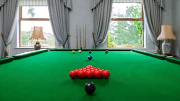 Una mesa de billar con bolas dispuestas en forma de triángulo, una ventana con cortinas y una lámpara al fondo.