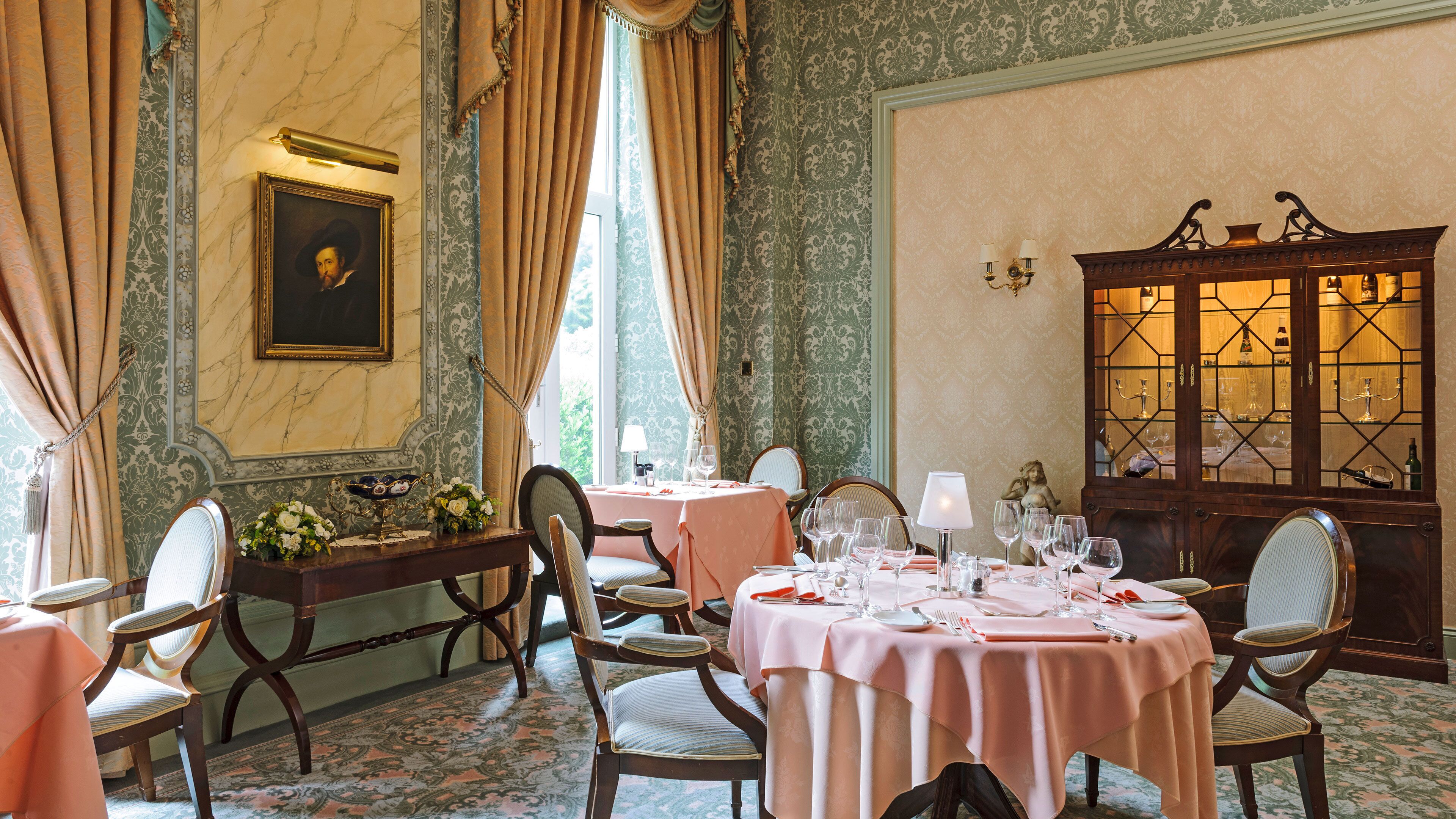 Un comedor con mesas redondas dispuestas para una comida, sillas, un aparador decorativo con puertas de cristal y un cuadro en la pared.