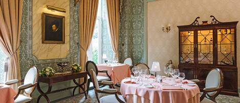 A dining room with round tables set for a meal, chairs, a decorative cabinet with glass doors, and a painting on the wall.