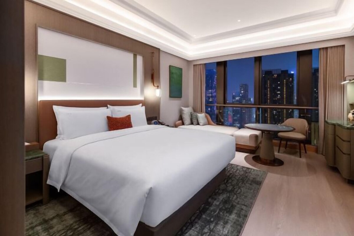 A hotel room with a large bed, a small table, a chair, and a view of the city at night.