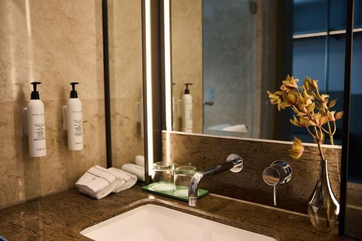A bathroom with a sink, mirror, toiletries, and a vase with flowers.