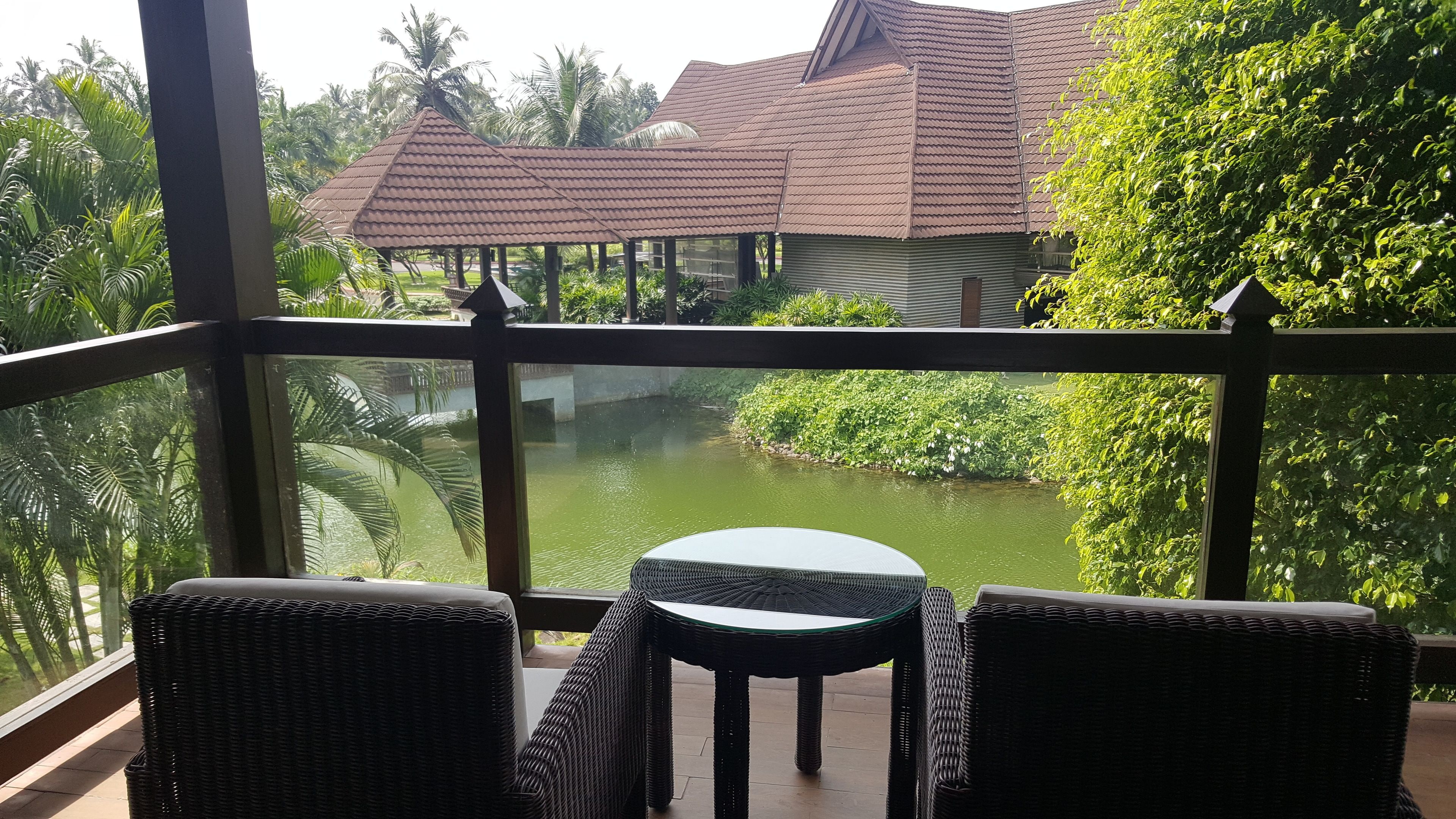 Ein Balkon mit Korbmöbeln, der auf einen Teich und ein Gebäude mit Ziegeldach blickt.