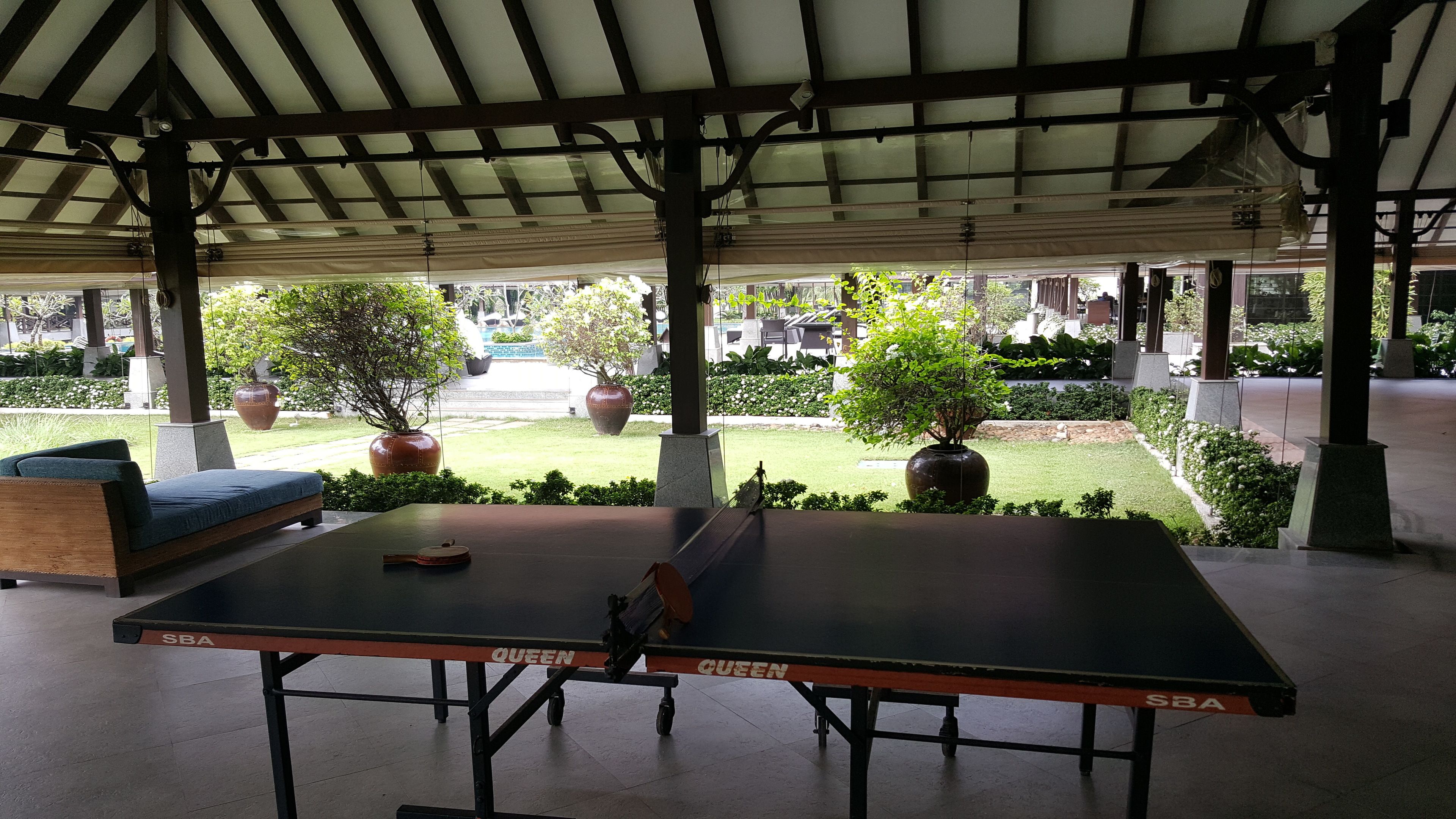 An outdoor table tennis table with a canopy overhead, surrounded by greenery and potted plants.