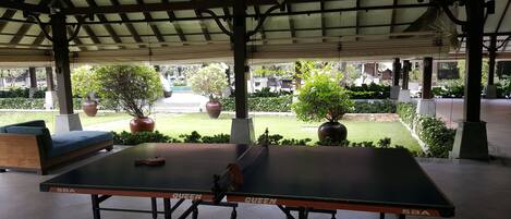An outdoor table tennis table with a canopy overhead, surrounded by greenery and potted plants.