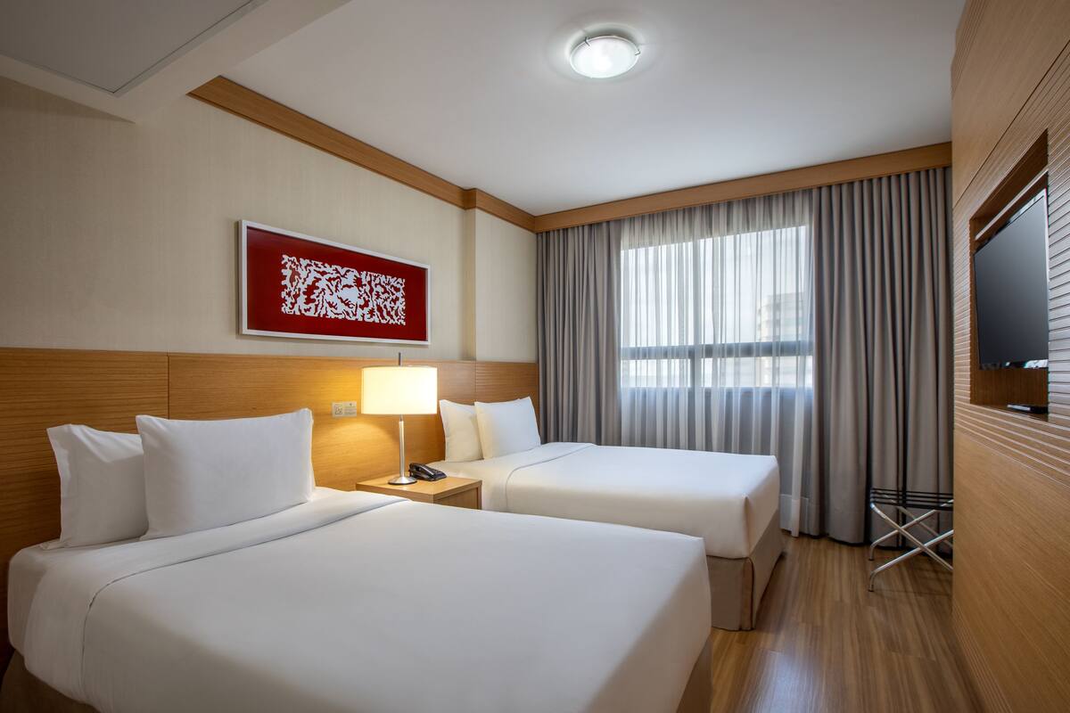 A hotel room with two beds, a bedside lamp, a flat-screen TV, a window with curtains, and a wall-mounted artwork.