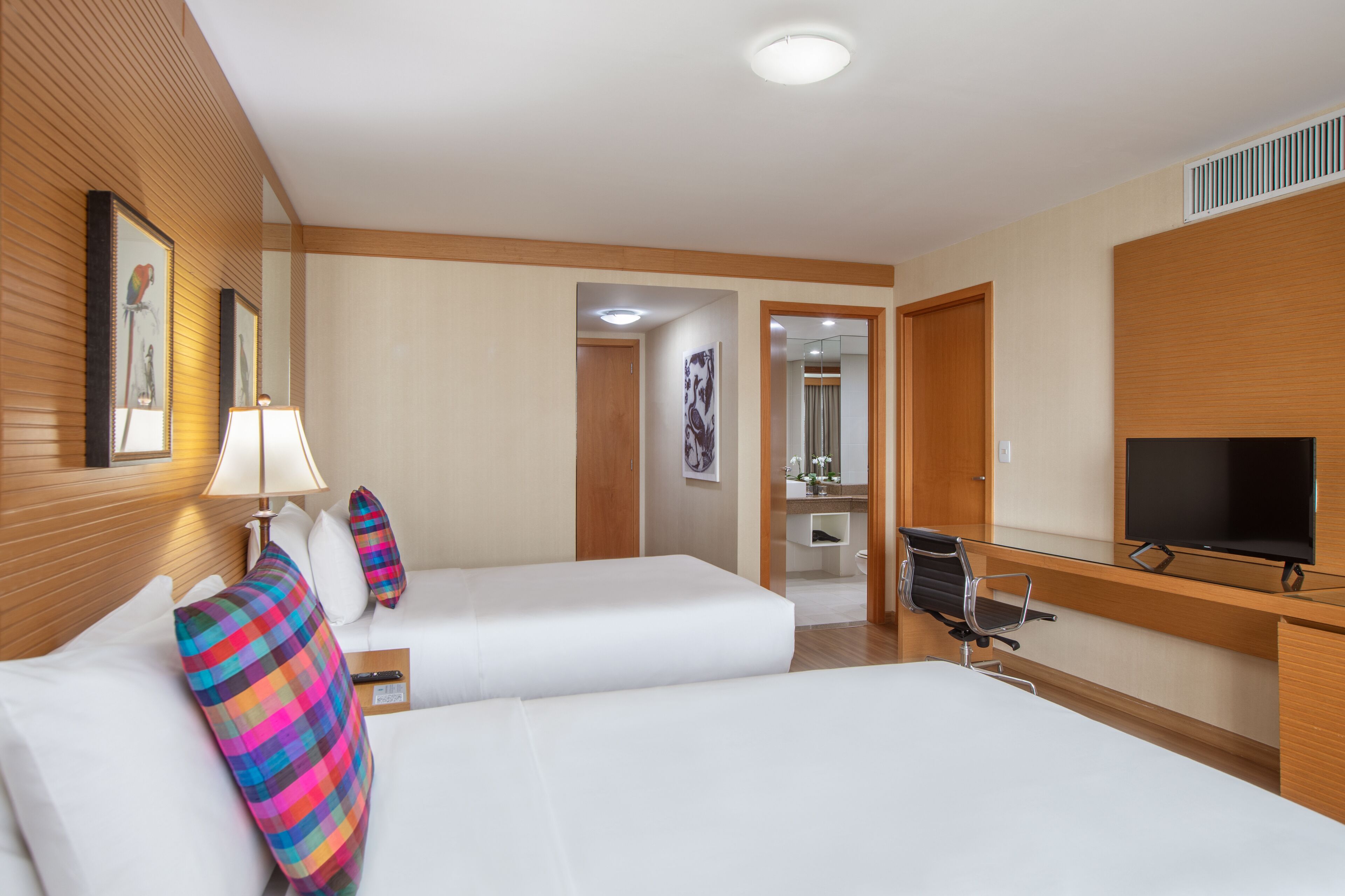 Habitación de hotel con dos camas, un televisor, un escritorio y un baño visible a través de una puerta abierta.