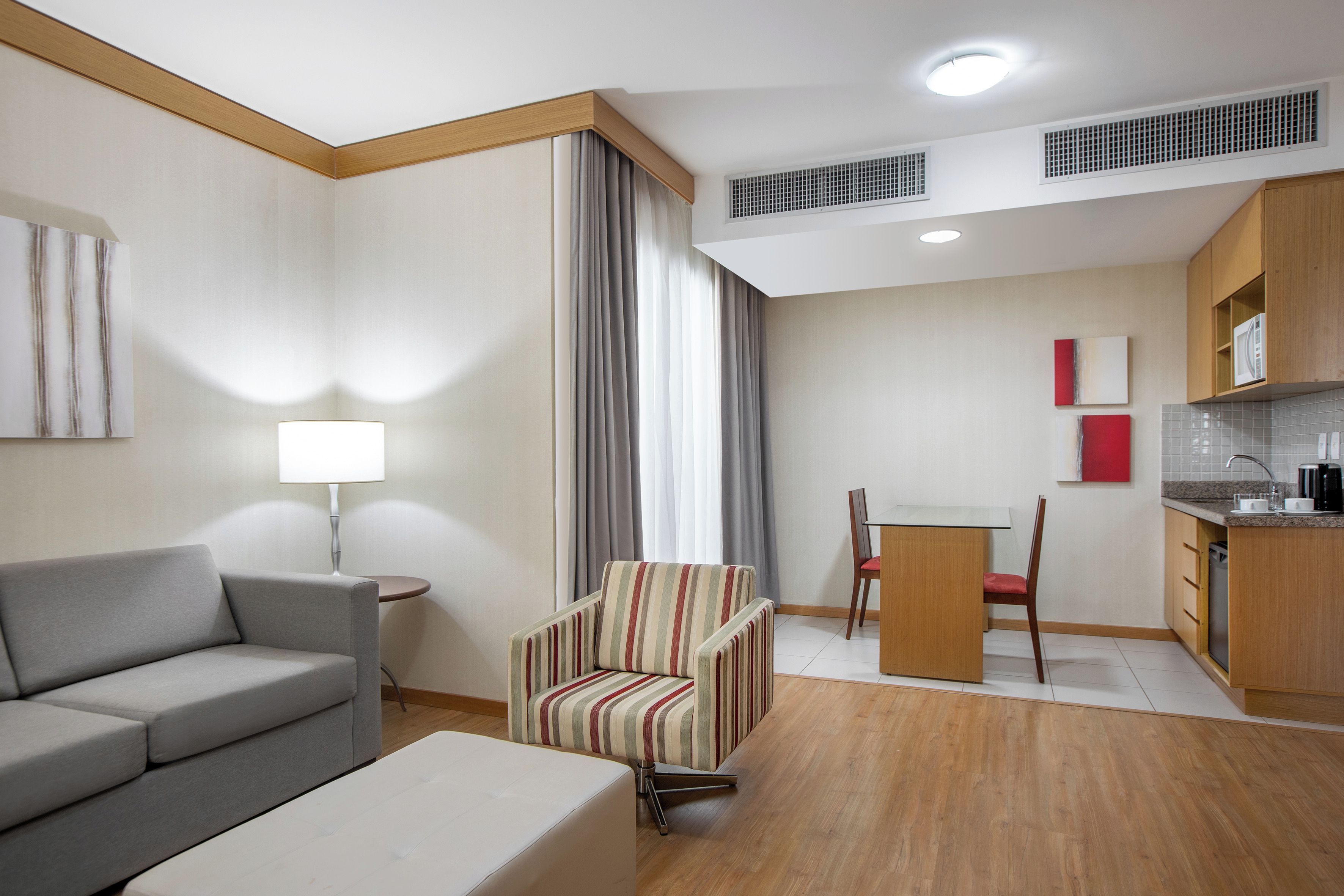 Habitación de hotel moderna con sofá gris, un sillón a rayas, una mesita con lámpara, zona de comedor con una silla y una kitchenette con microondas.