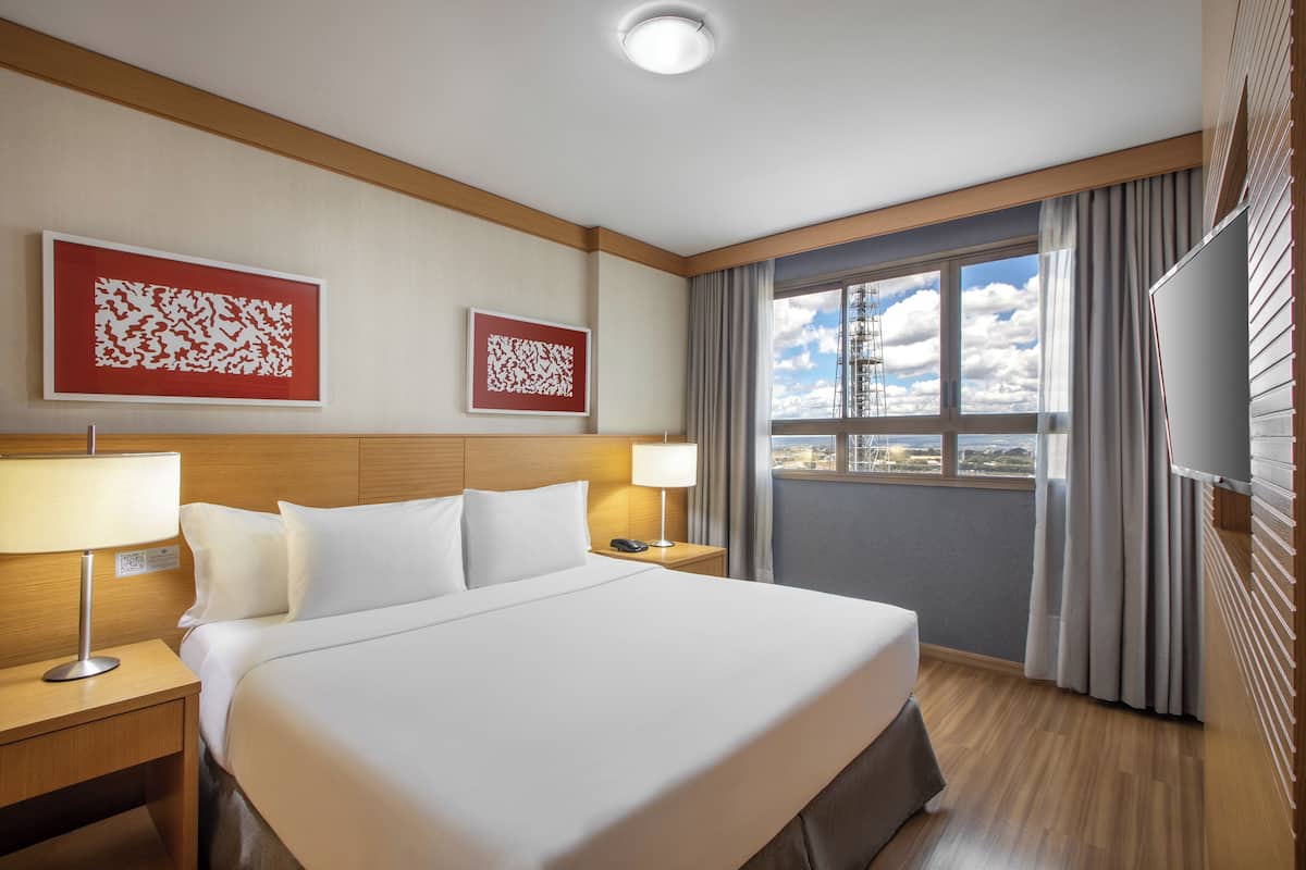 A hotel room with a large bed, two bedside tables with lamps, a flat-screen TV, and a window with curtains.