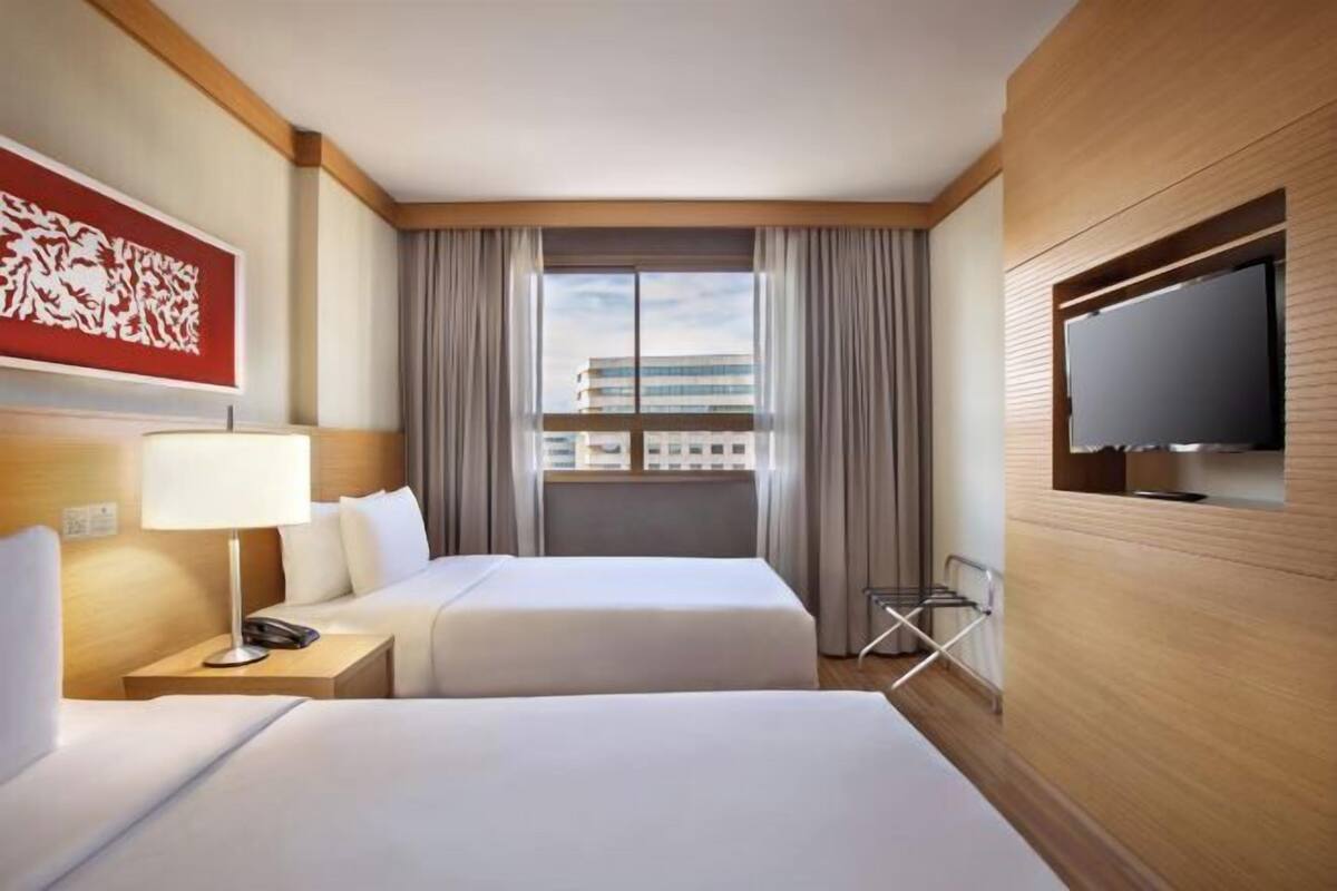 A hotel room with two beds, a TV, a window with curtains, a lamp, and a wall-mounted artwork.