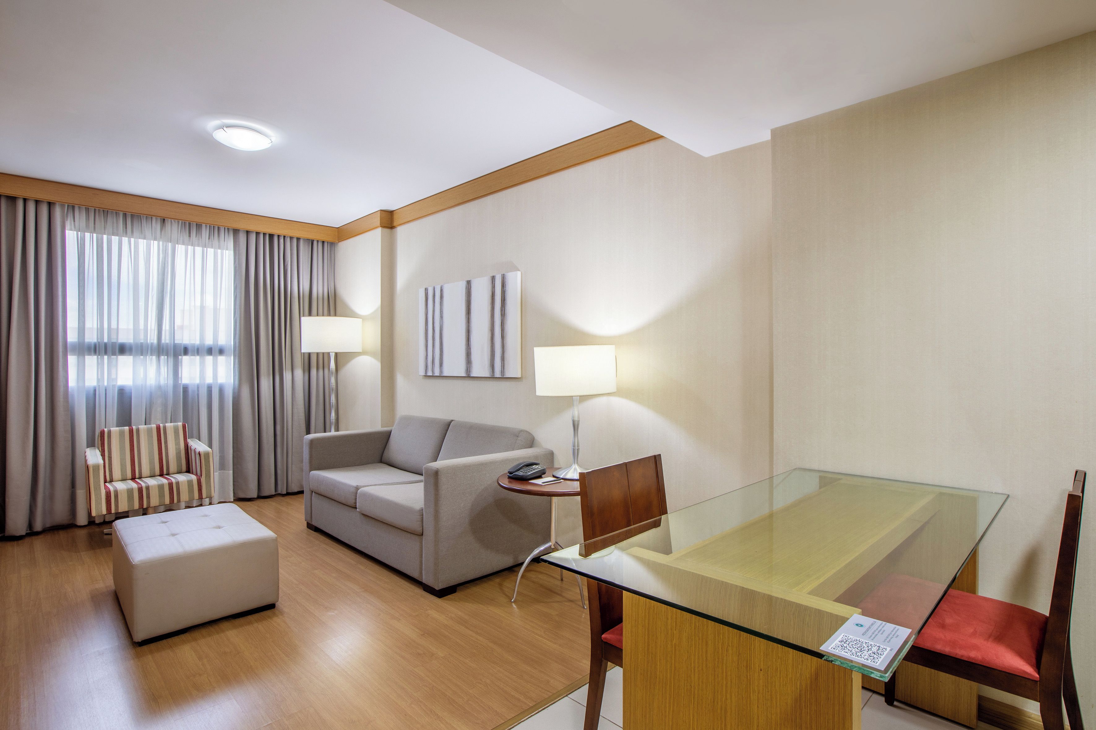 Habitación de hotel con mesa de comedor de vidrio, sofá, una silla, lámpara y un ventanal con cortinas.