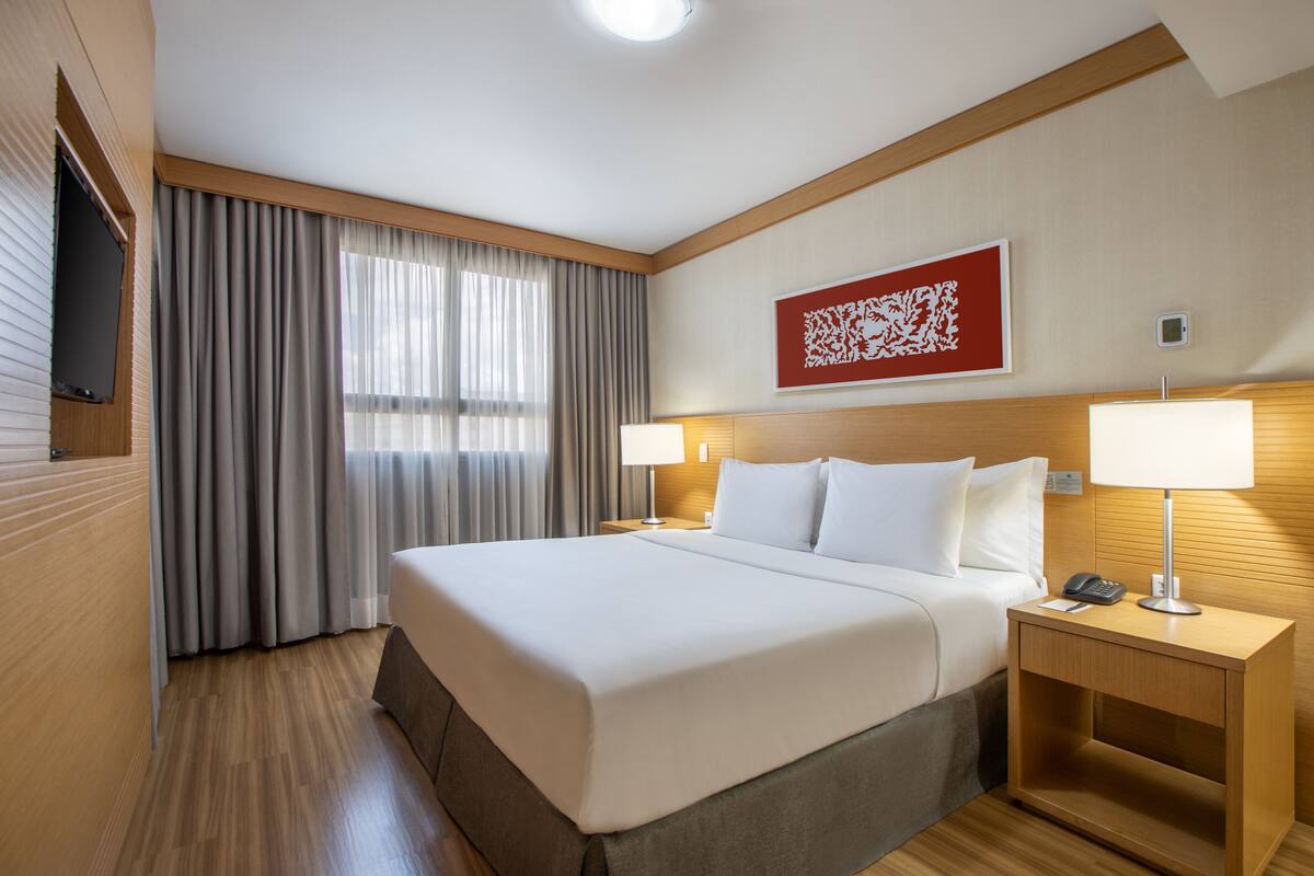 A hotel room with a large bed, two bedside tables with lamps, a flat-screen TV mounted on the wall, and a window with curtains.