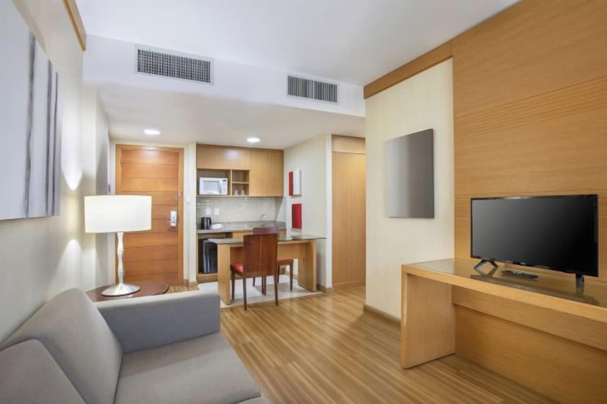 Una habitación de hotel moderna con sofá, televisor, escritorio de madera y una kitchenette al fondo.