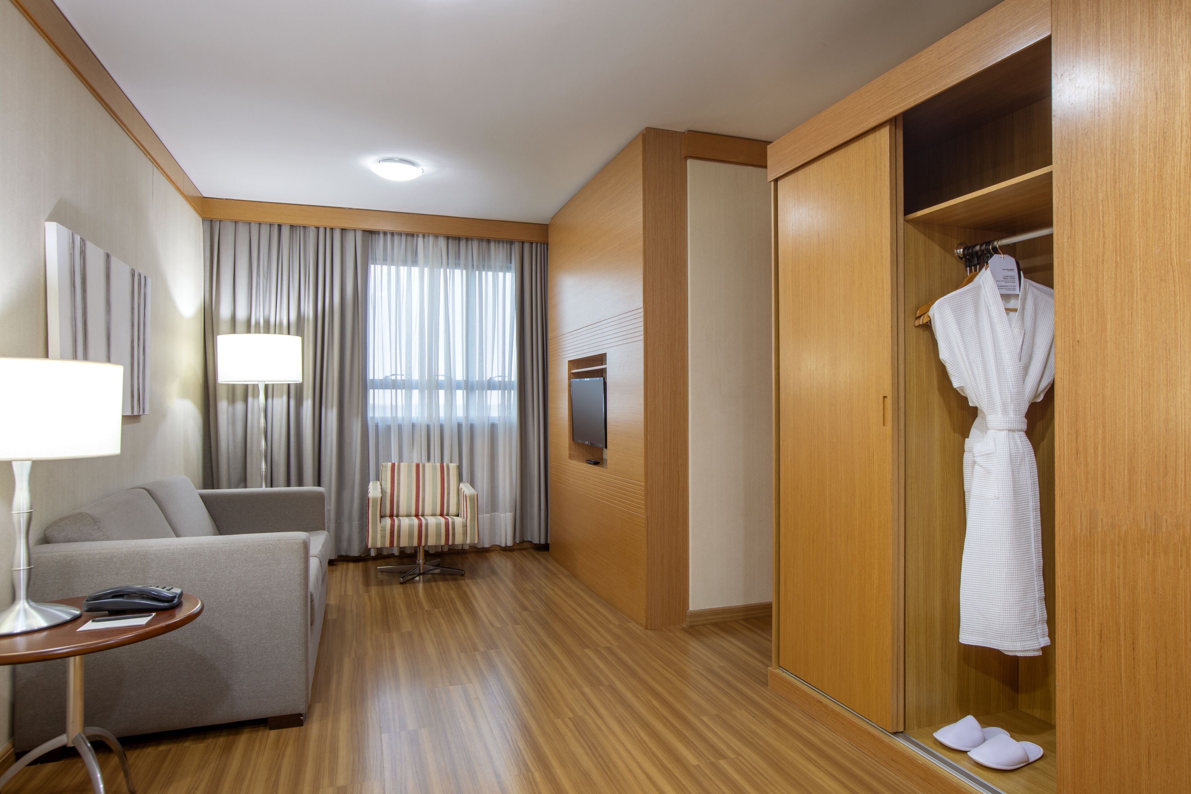 A hotel room with a wooden floor, a sofa, a table with a lamp, a TV, a wardrobe, and a robe hanging inside.