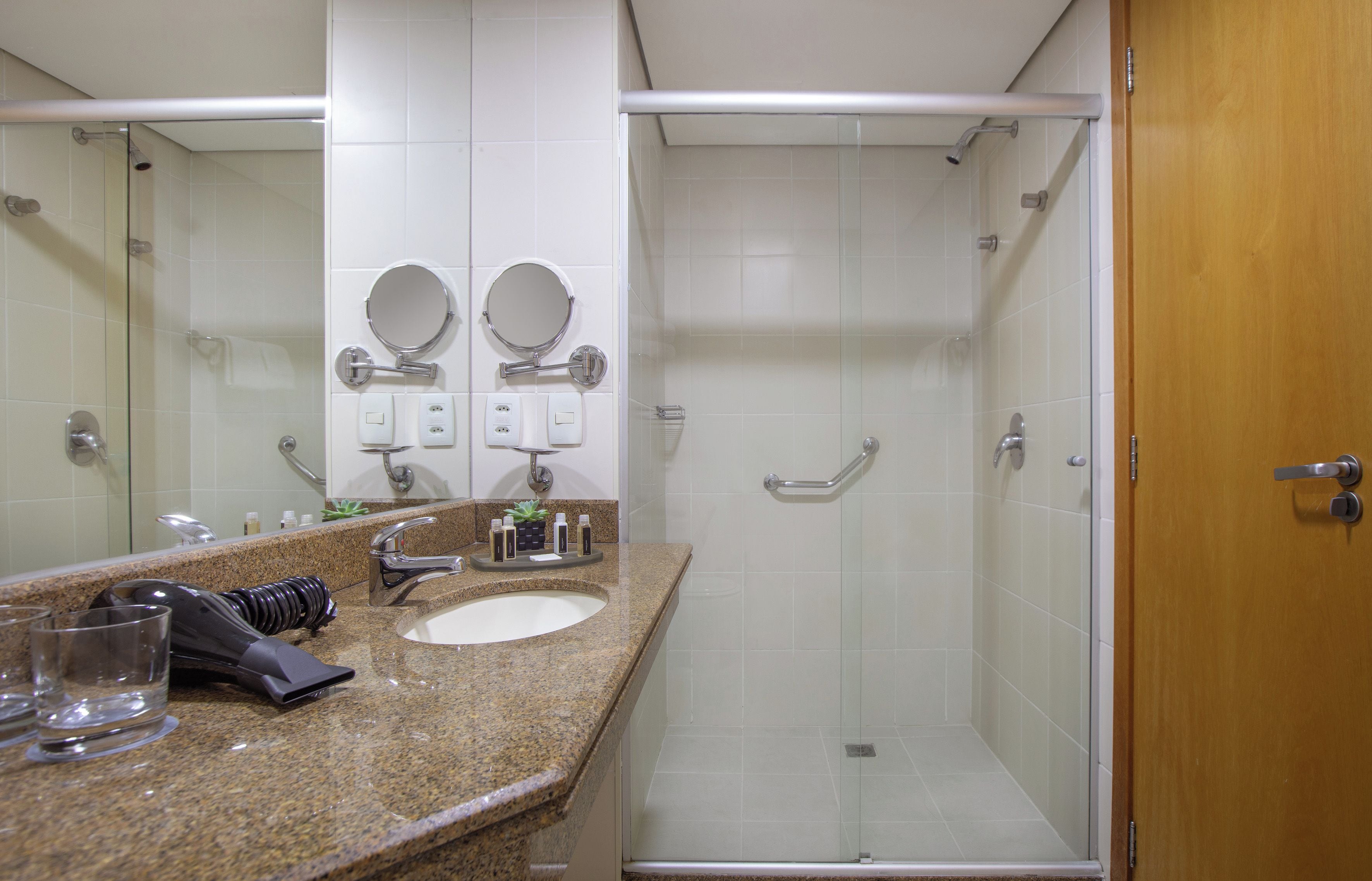 A bathroom with a granite countertop, two sinks, a shower with a glass door, and a wooden door.