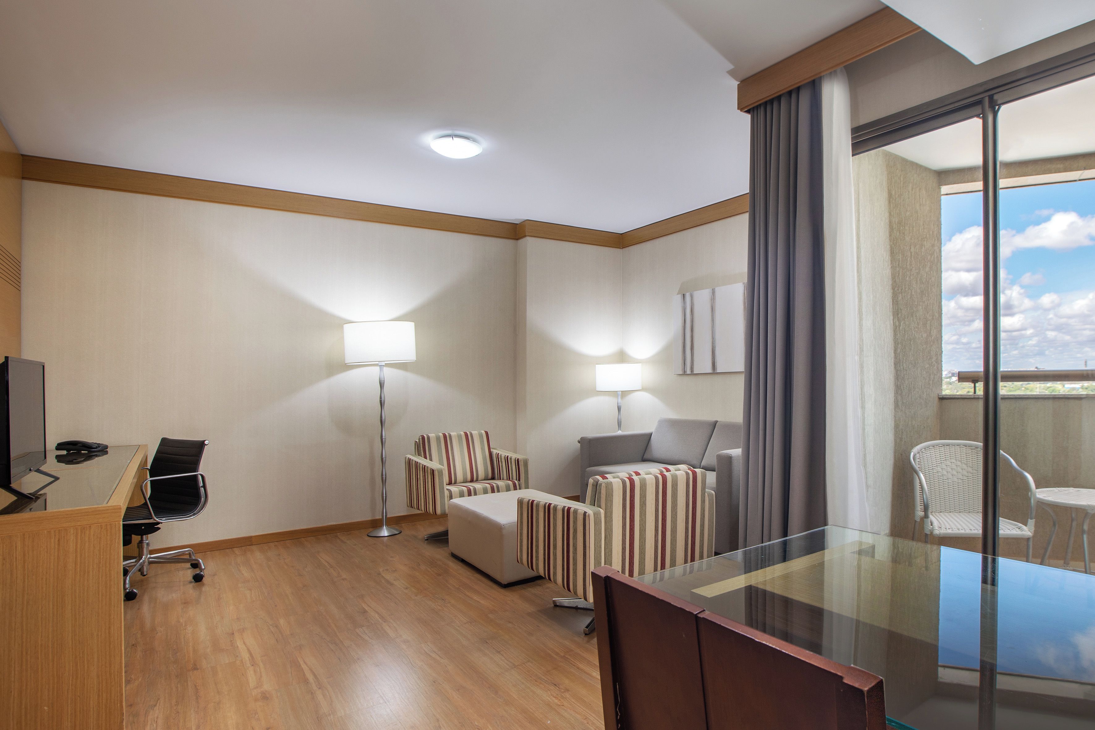 Een hotelkamer met een bureau, stoel, twee gestreepte fauteuils, een bank en een glazen bijzettafeltje, plus een vloerlamp.