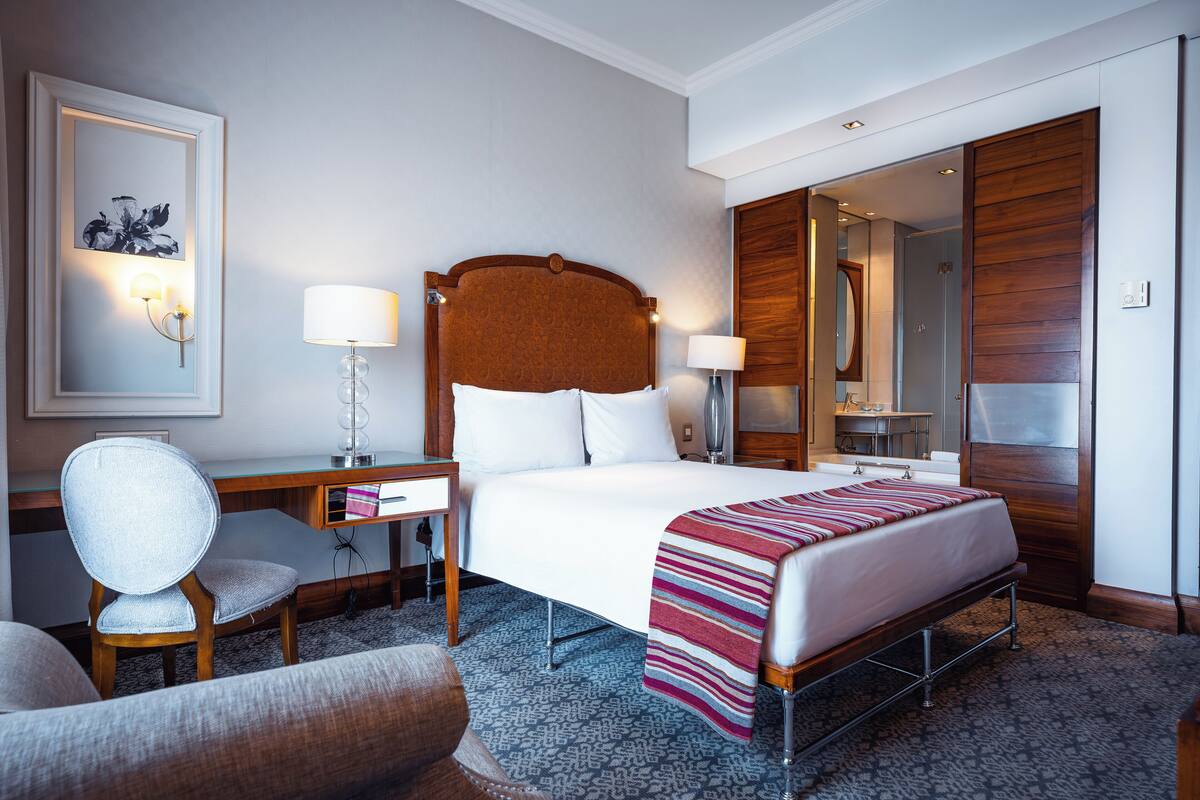 Habitación de hotel con una cama grande, un escritorio con una silla, una lámpara de escritorio, una mesita de noche, un espejo y un armario.
