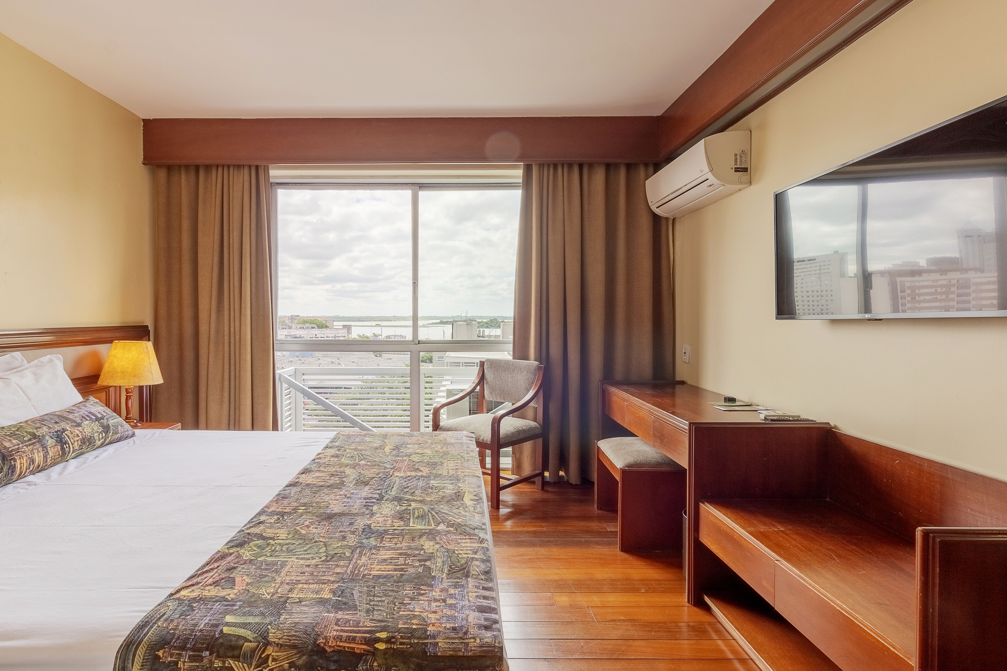 Une chambre d’hôtel avec un grand lit, un bureau, une chaise, un banc, une télévision et une fenêtre donnant sur des immeubles.