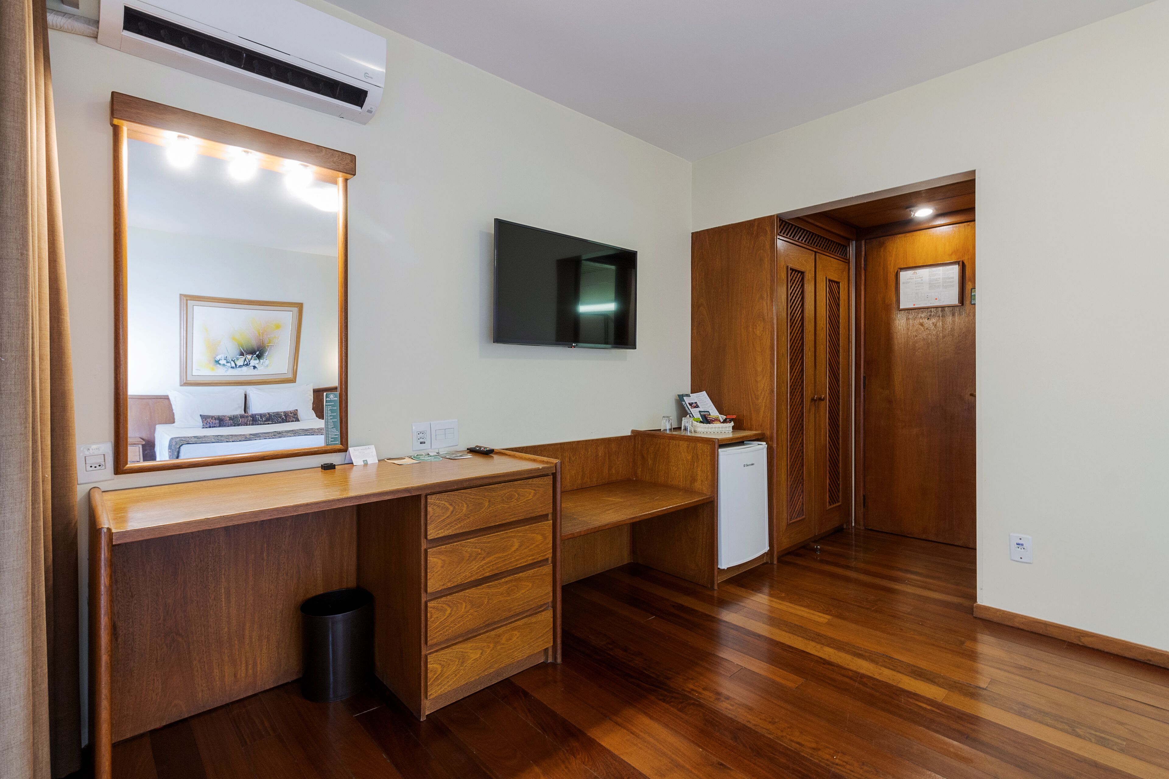 A hotel room with a wooden desk, a large mirror with lights, a flat-screen TV, a small refrigerator, and a wooden wardrobe.