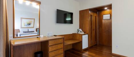A hotel room with a wooden desk, a large mirror with lights, a flat-screen TV, a small refrigerator, and a wooden wardrobe.