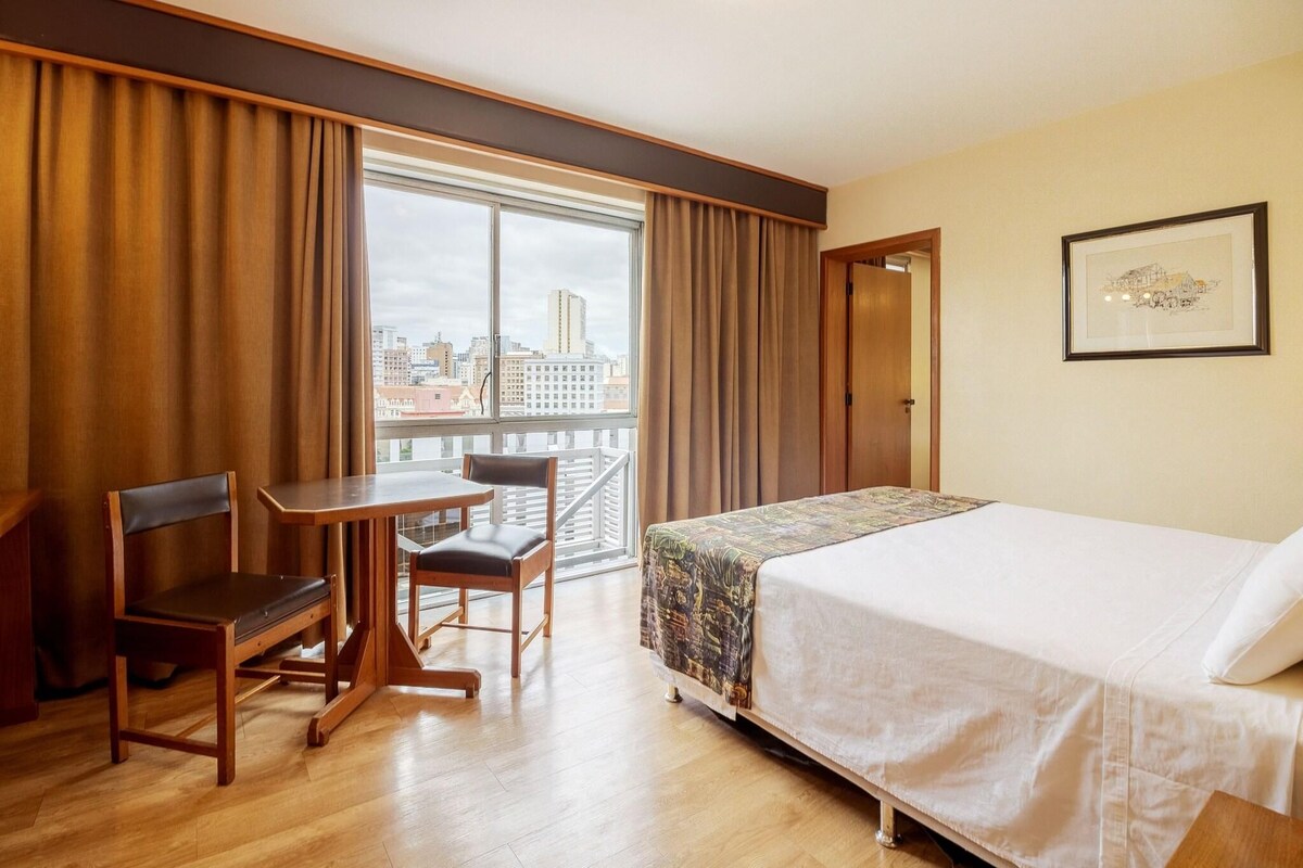 A hotel room with a bed, a desk, and a chair. There is a window with curtains, a door, and a framed picture on the wall.