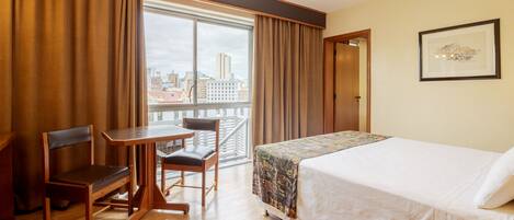 Habitación de hotel con cama, escritorio, dos sillas, ventana con cortinas e imagen en el marco en la pared.