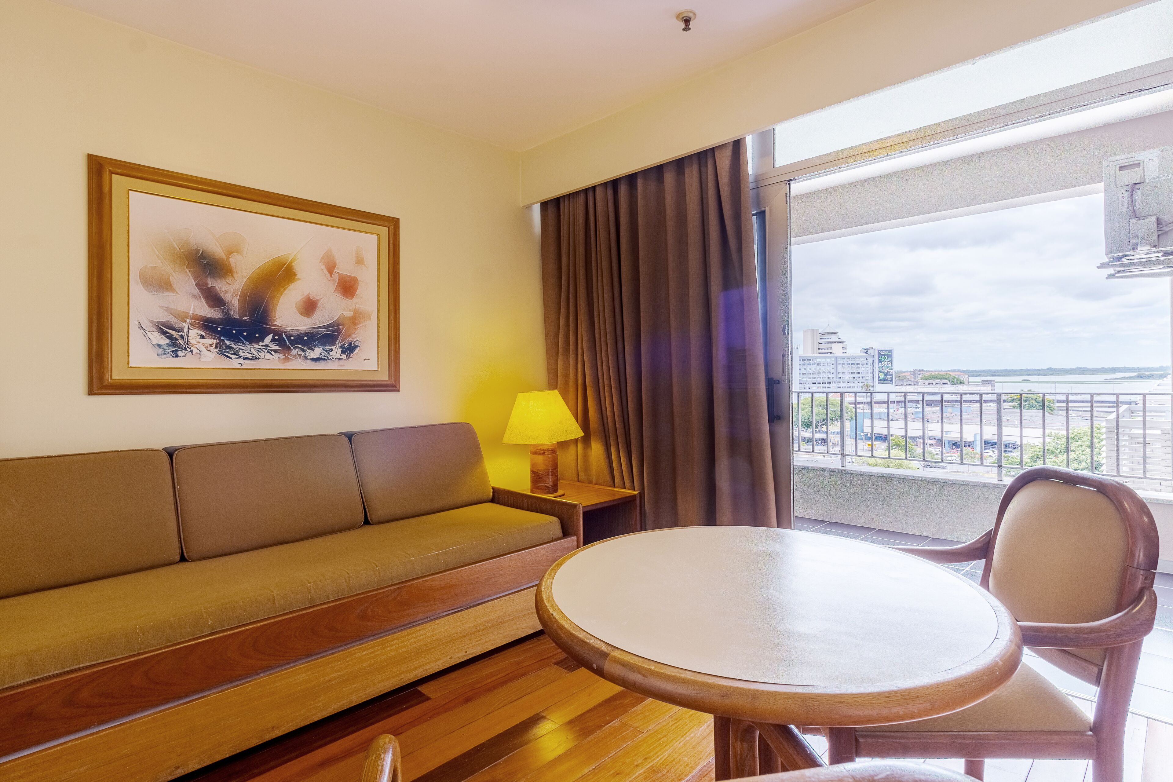 Una habitación con piso de madera, un sofá beige, una mesa redonda, una silla, una lámpara, un cuadro abstracto enmarco y vistas de la ciudad a través de una ventana con cortinas.