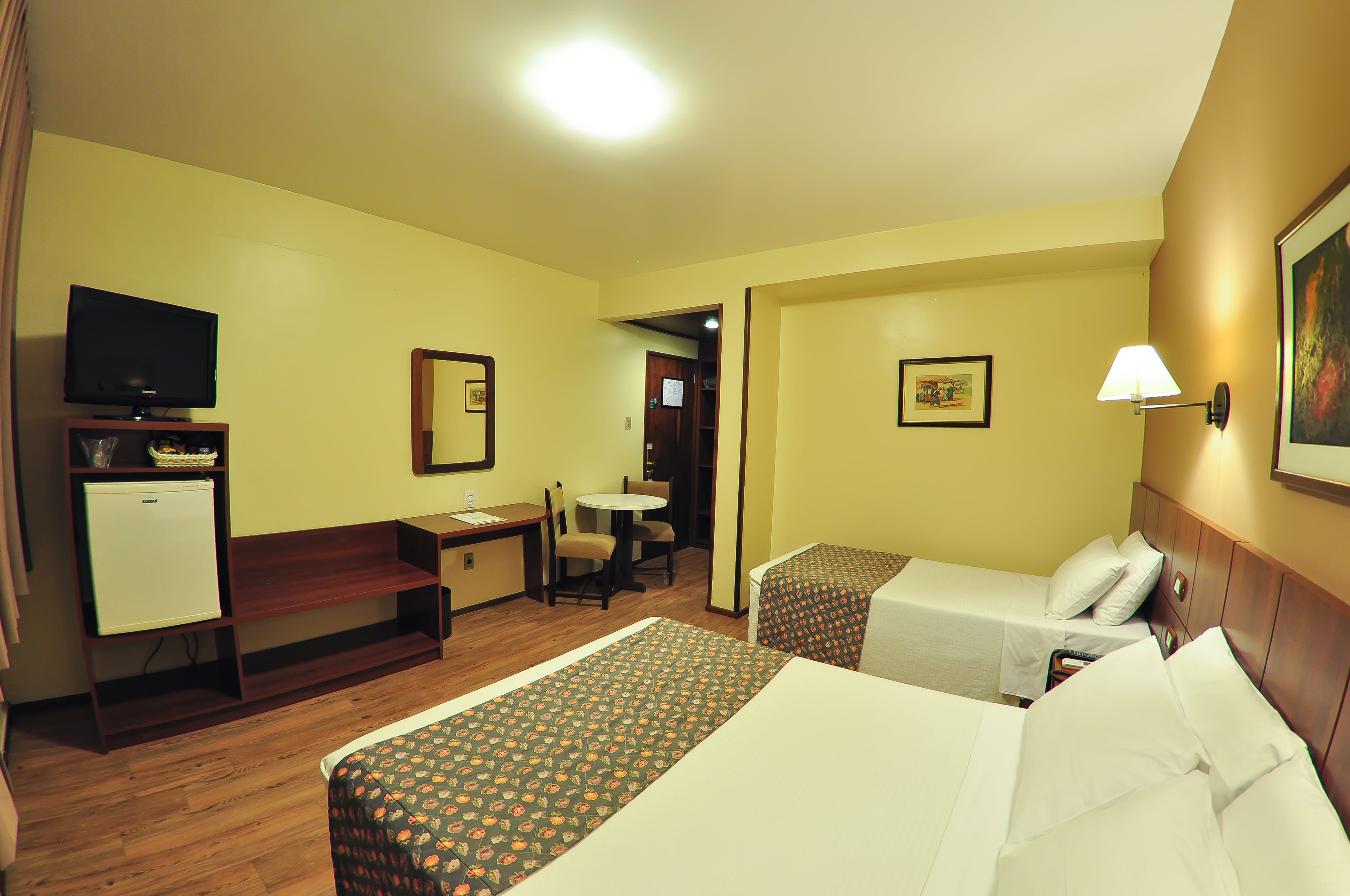 Habitación de hotel con dos camas individuales, un escritorio con silla, televisión y un refrigerador pequeño.