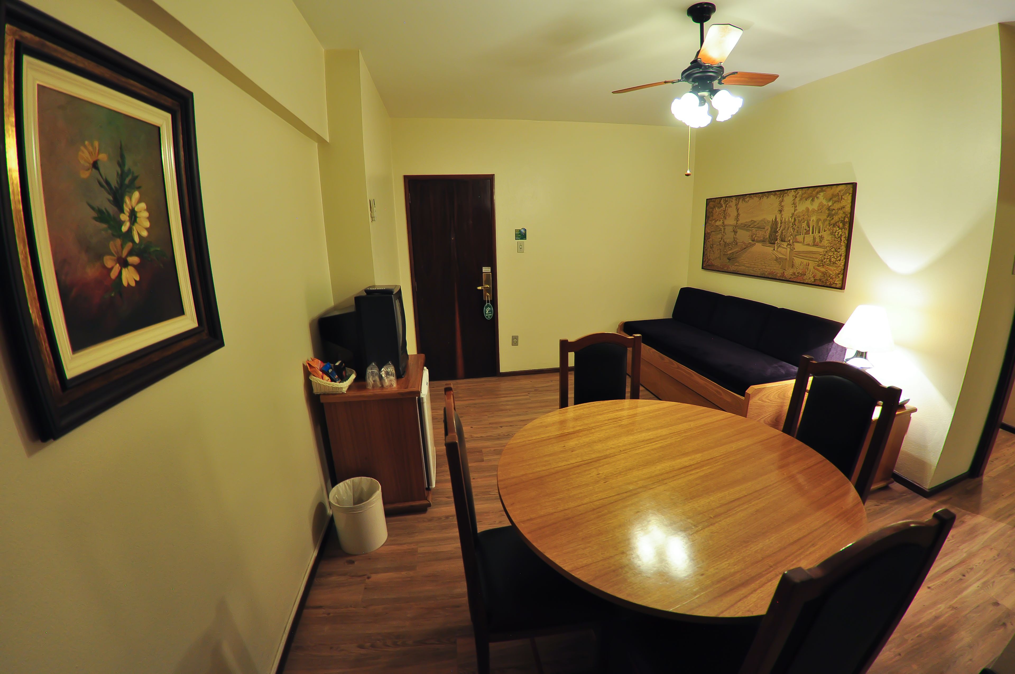 A dining area with a round wooden table, chairs, a sofa, a ceiling fan, and a framed painting on the wall.