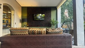 A modern living room with a large sofa, patterned cushions, a flat-screen TV, and a view of greenery outside. - Ritter Hotéis (Porto Alegre)