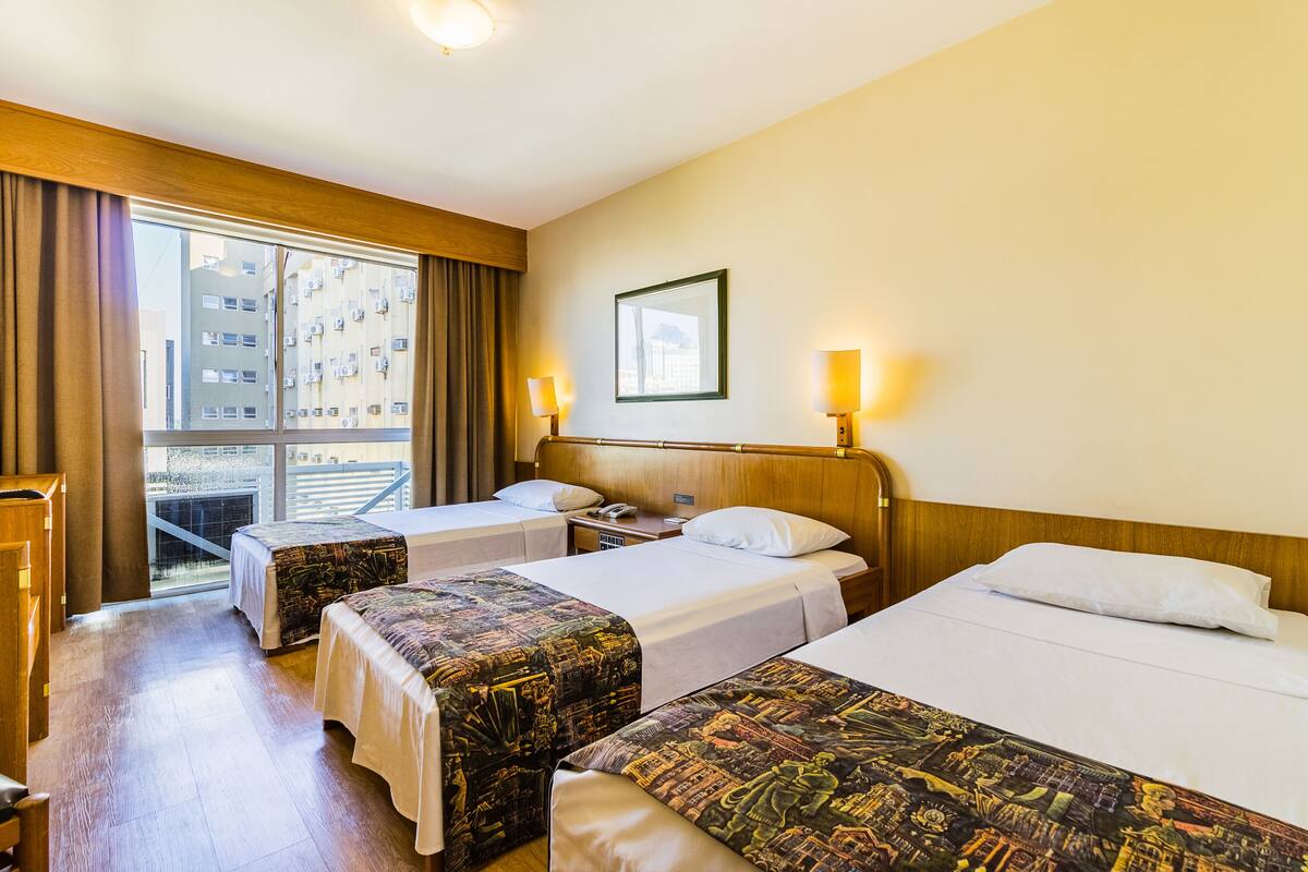 A hotel room with two beds, a wooden headboard, a large window with curtains, and a view of buildings outside.