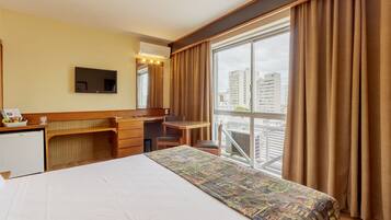 Quarto de hotel com cama, escrivaninha, cadeira, televisão e janela com cortinas.