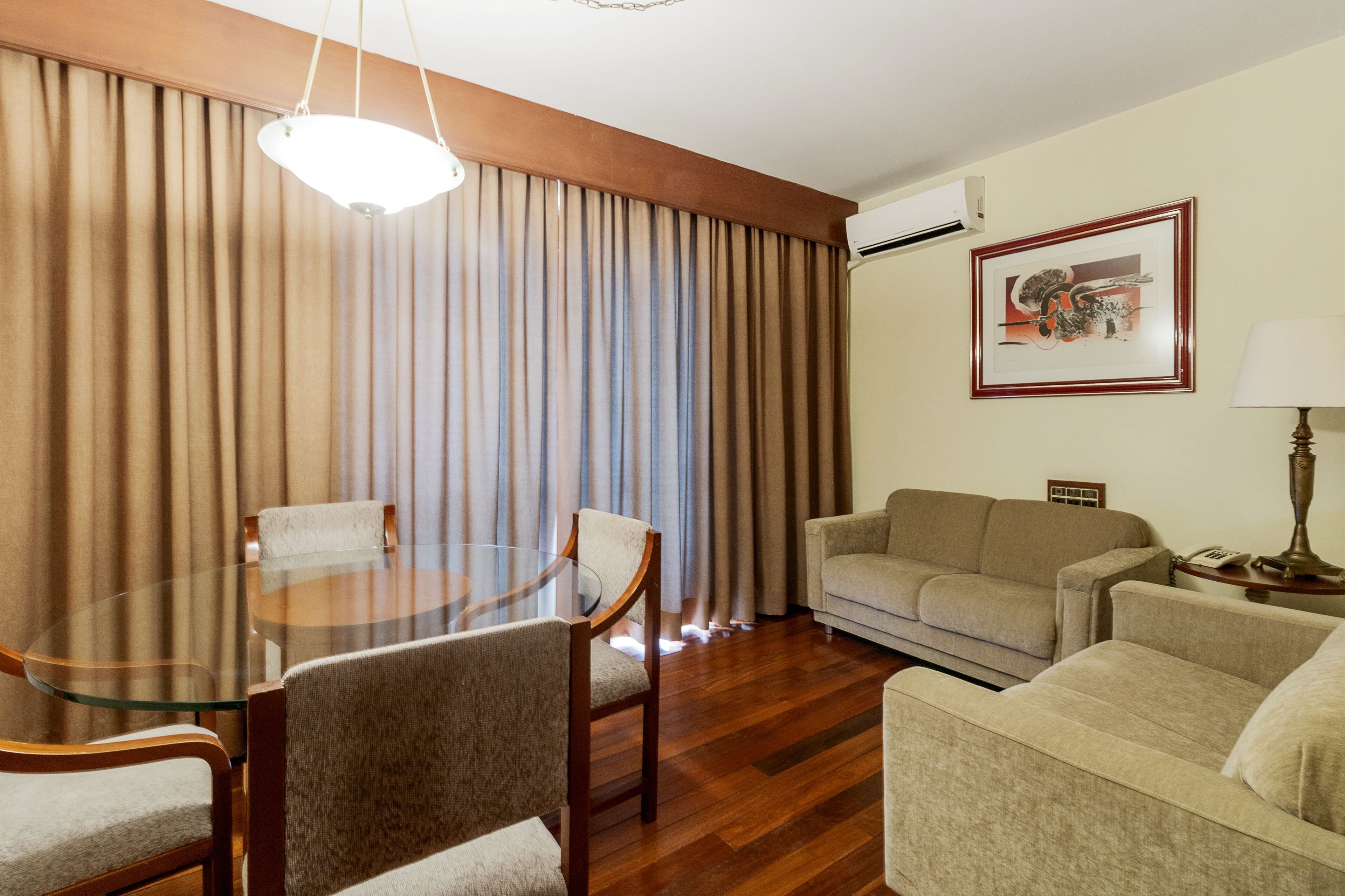 A hotel room with a round glass table, two armchairs, a sofa, a lamp, and a framed picture on the wall.