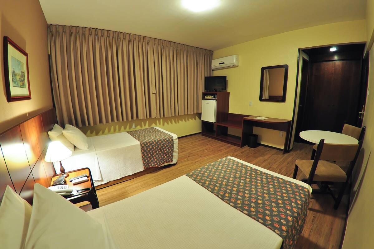 A hotel room with two beds, a desk, a chair, a TV, and an air conditioning unit.