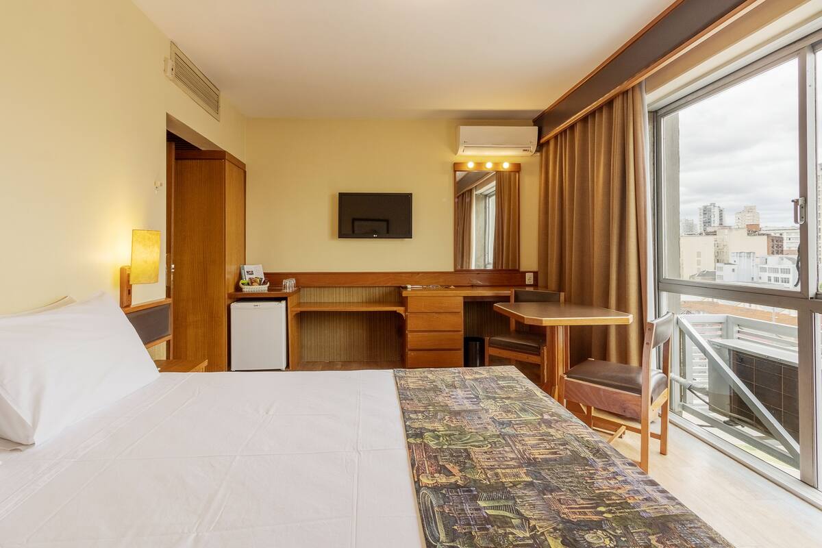 A hotel room with a bed, a desk, a chair, a small refrigerator, and a large window offering a city view.