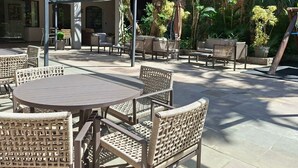An outdoor patio area with round tables and wicker chairs, covered by a glass roof, surrounded by greenery and a building in the background. - Ritter Hotéis (Porto Alegre)
