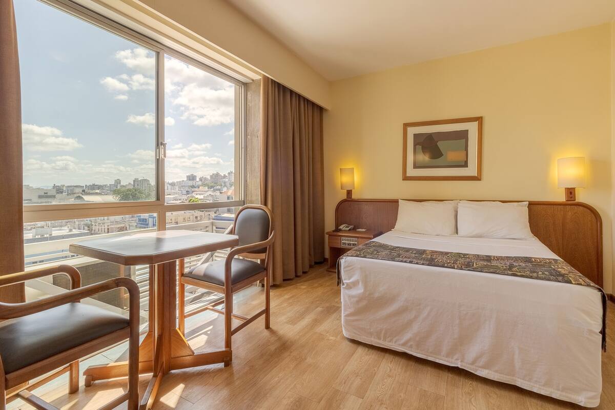 A hotel room with a bed, a desk, a chair, a nightstand, a lamp, and a window with a city view.