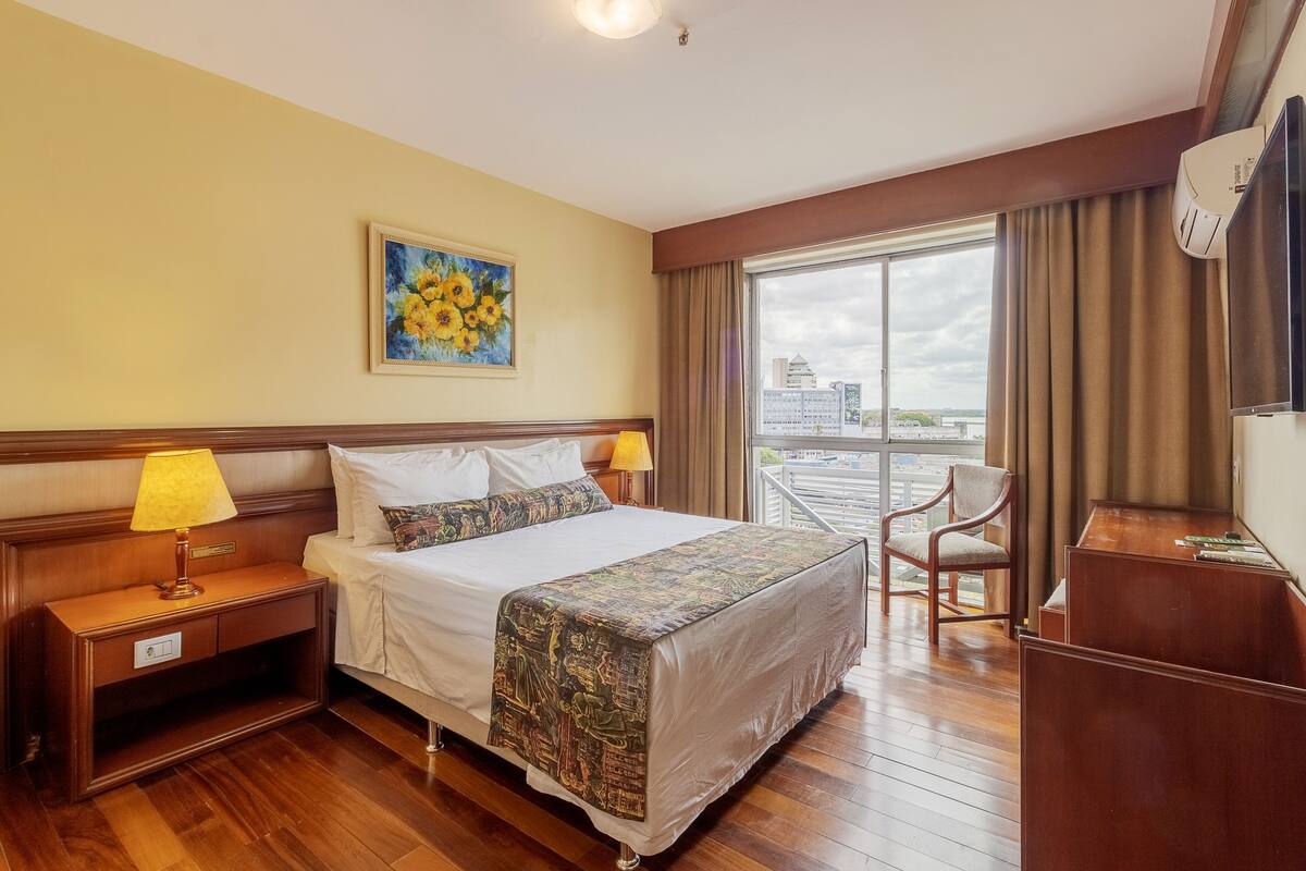 A hotel room with a bed, a nightstand, a desk, a chair, and a large window offering a city view.