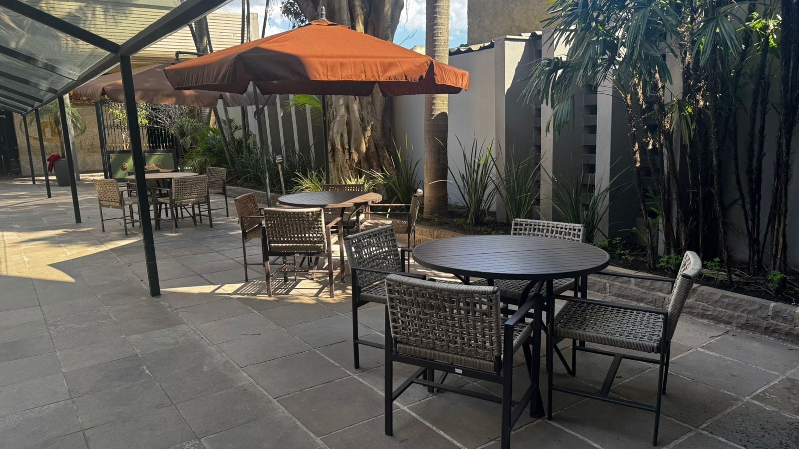 An outdoor patio with round tables and wicker chairs, shaded by a large umbrella.