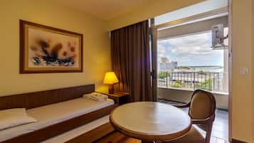 Quarto de hotel com cama, mesa, cadeira, abajur, pintura e varanda com vista para a cidade.