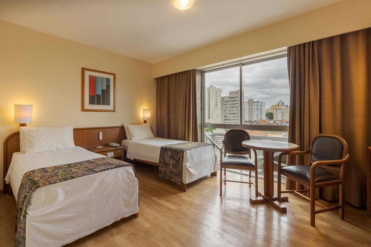 A hotel room with two beds, a small table with chairs, a view of the city through large windows, and a painting on the wall.